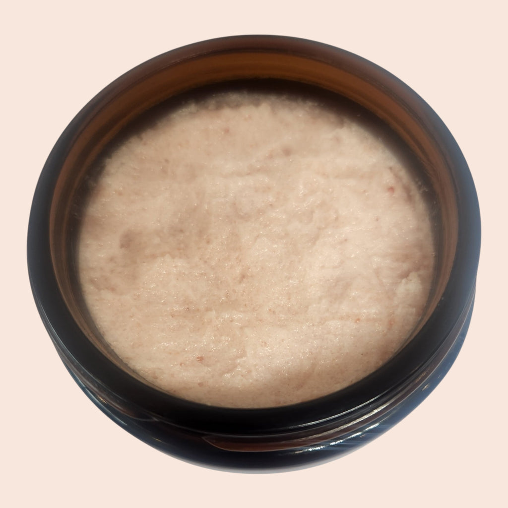 A top-down view of a round dark amber glass jar filled with a light beige textured cosmetic cream or scrub, set against a solid pale peach background.
