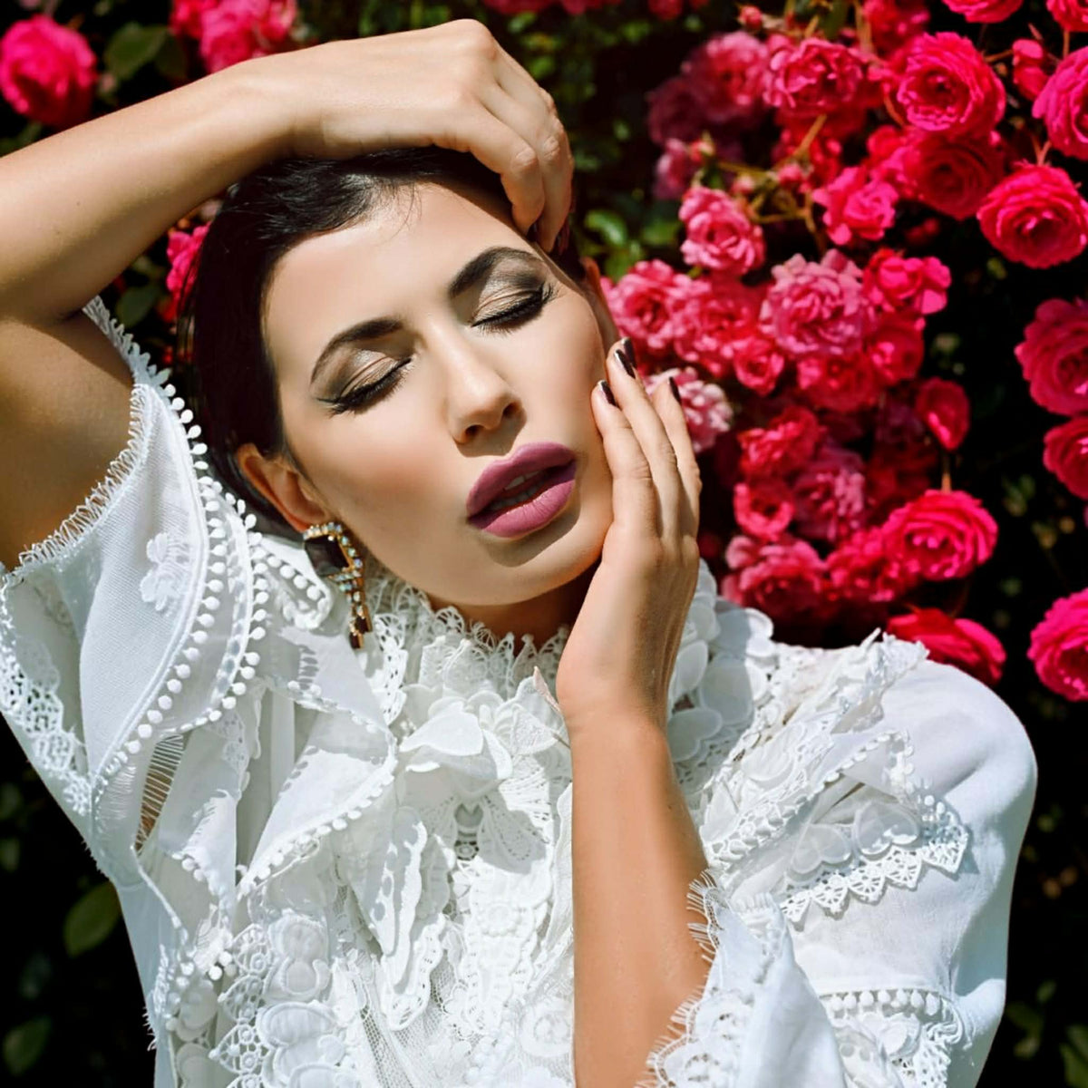A woman in a white lace blouse poses outdoors against a dense background of bright pink roses.