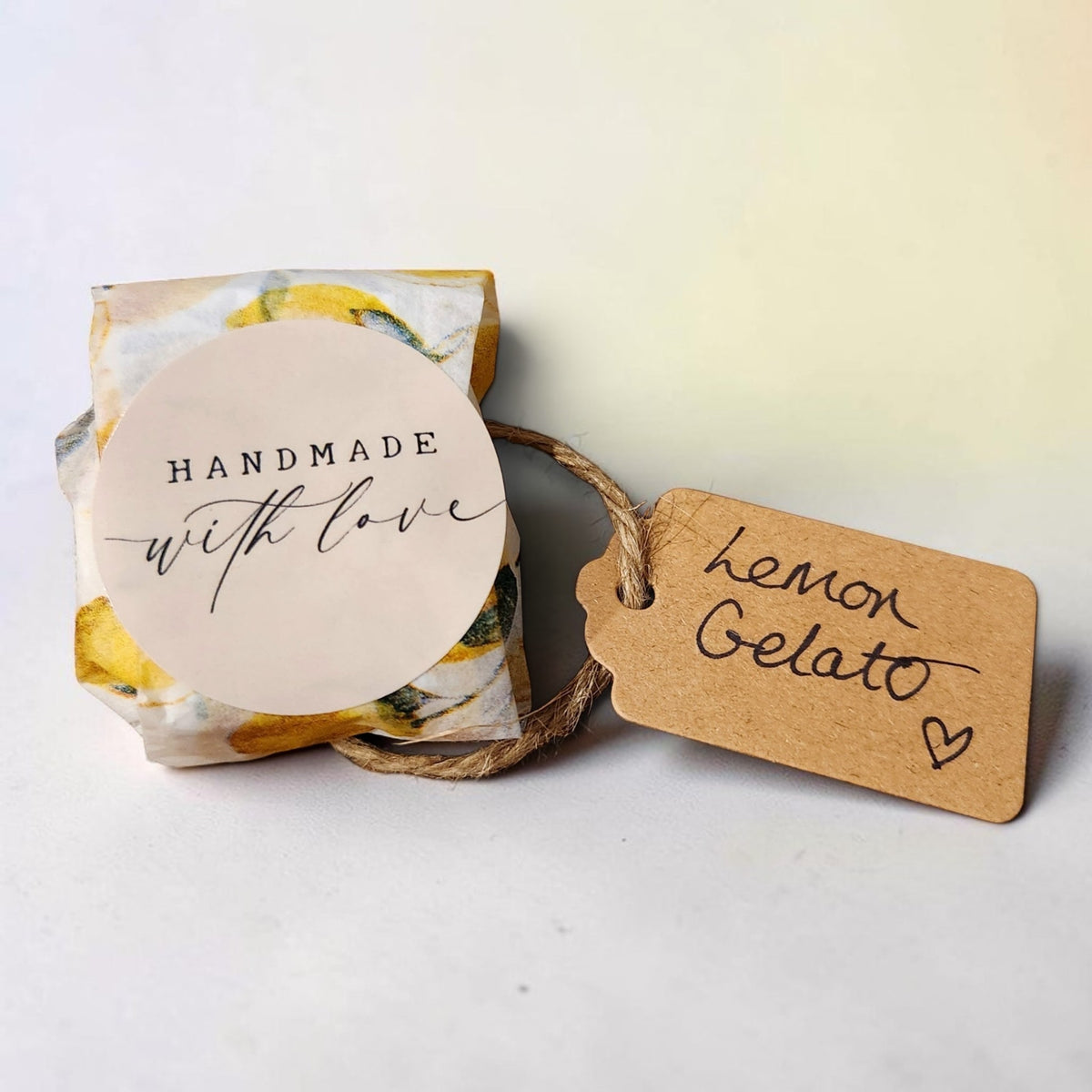 A small wrapped gift box with a round cream paper label reading 'HANDMADE with love' and a kraft paper hang tag tied with twine that says 'Lemon Gelato' in black script, photographed on a light neutral background.