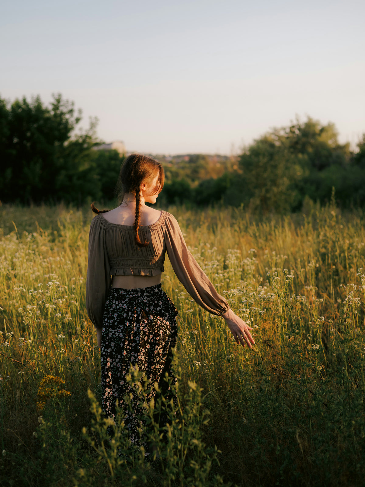 A woman stands in a sunlit grassy field, seen from behind, wearing a taupe long-sleeve crop top and a black floral maxi skirt. The background features a soft-focus landscape of trees and a clear sky during golden hour.